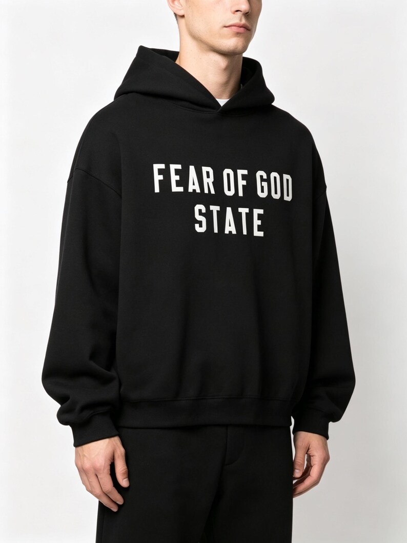 May include: Black hooded sweatshirt with the text "FEAR OF GOD STATE" in white block letters. The sweatshirt has long sleeves and a relaxed fit. The garment is made of a soft material.