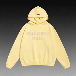 May include: A pale yellow hoodie with the text "FEAR OF GOD STATE" printed on the front. The hoodie has a hood with a small, dark-colored tag at the top. The garment is made of a soft material and has long sleeves.