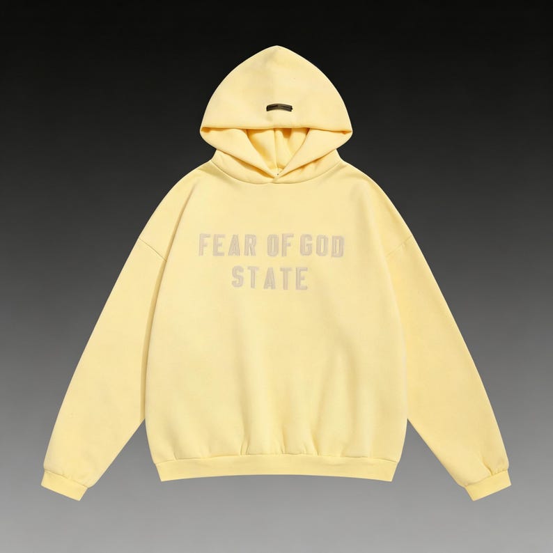 May include: A pale yellow hoodie with the text "FEAR OF GOD STATE" printed on the front. The hoodie has a hood with a small, dark-colored tag at the top. The garment is made of a soft material and has long sleeves.