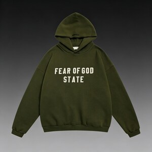 May include: An olive green hoodie with the text "FEAR OF GOD STATE" in white block letters across the chest. The hoodie has a drawstring hood and long sleeves. The background is a gradient of dark gray and black.