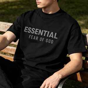 May include: Black t-shirt with the text "ESSENTIAL FEAR OF GOD" in gray. The shirt is short-sleeved and has a crew neck. The person is sitting on a wooden bench outside.