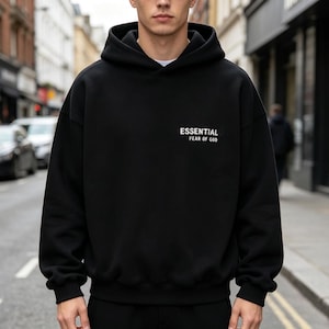 Op de afbeelding: Zwarte hoodie met de tekst "ESSENTIAL FEAR OF GOD" in het wit. De hoodie heeft een capuchon met trekkoord en een relaxte pasvorm. De persoon staat op een stadsstraat.