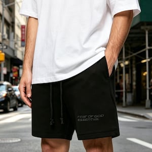 Pode incluir: Calções pretos com cintura com cordão e o texto "FEAR OF GOD ESSENTIAL" em cinza. Os calções são combinados com uma camiseta branca. A imagem é tirada ao ar livre numa rua da cidade.