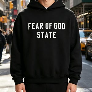 May include: Black hooded sweatshirt with the text "FEAR OF GOD STATE" in white block letters. The sweatshirt has a drawstring hood and long sleeves. The person is standing in front of a city street.