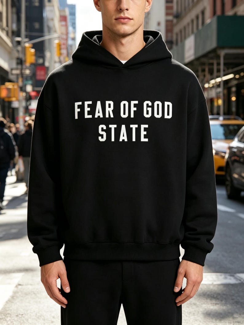May include: Black hooded sweatshirt with the text "FEAR OF GOD STATE" in white block letters. The sweatshirt has a drawstring hood and long sleeves. The person is standing in front of a city street.