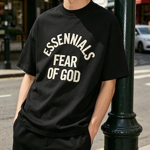 May include: Black t-shirt with the text "ESSENTIALS FEAR OF GOD" in white, arched above and below. The shirt is short-sleeved and appears to be made of a soft material. The person is wearing black pants and is leaning against a dark green pole.