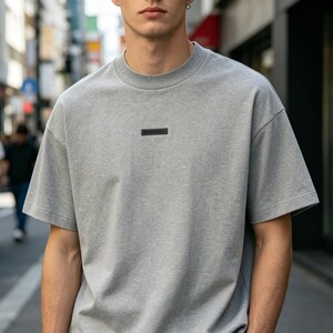 May include: A light gray t-shirt with a crew neck and short sleeves. A small, dark rectangular patch is centered on the chest. The shirt has a relaxed fit and is made of a soft material.