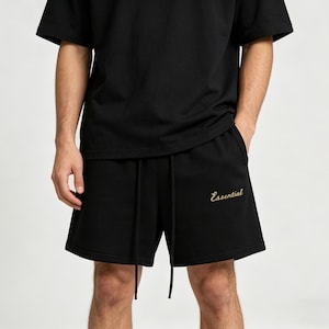 Op de afbeelding: Zwarte shorts met een tailleband met trekkoord en het woord "Essential" geborduurd in goud. De shorts maken deel uit van een bijpassende zwarte t-shirtset.