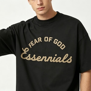 May include: Black short-sleeved t-shirt with the text "FEAR OF GOD Essentials" in a curved, gold-colored font. The shirt has a crew neck and a relaxed fit, suitable for casual wear.