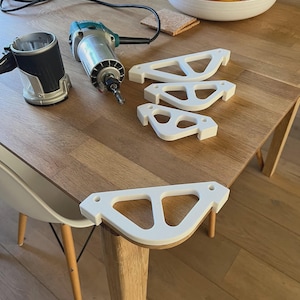 Op de afbeelding: Een houtbewerkingsscène met een elektrische freesmachine en verschillende witte, gebogen beugels met een geometrisch ontwerp. De gereedschappen en componenten zijn gerangschikt op een houten tafel, wat wijst op een doe-het-zelfproject of meubelconstructie.