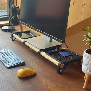 Op de afbeelding: Een houten monitorstandaard met een zwart frame en een zilveren bovenkant, met een computermonitor. Een draadloos toetsenbord en een gele muis liggen op het bureau. Een kleine plant in een pot is ook zichtbaar.