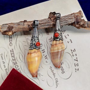 Può includere: Due pendenti composti da conchiglie coniche color marrone e beige con montature in metallo argentato decorate. Ogni cappuccio in metallo presenta un piccolo inserto rotondo in pietra rosso vivo e un occhiello superiore per l'aggancio.