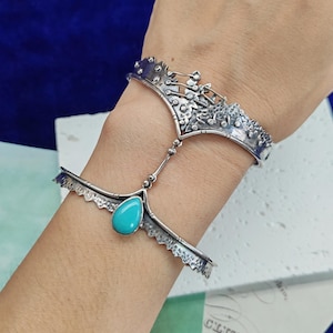 Peut inclure: Un bracelet esclave en argent composé d'une manchette en métal oxydé texturé reliée par une fine chaîne à une pièce de main assortie. La pièce de main est ornée d'une unique pierre turquoise lisse en forme de goutte au centre de la bande.