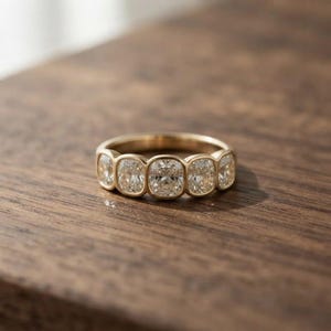 May include: A gold band ring featuring five cushion-cut clear stones set in a row. The stones are arranged horizontally across the top of the band, each secured within a smooth, rounded gold bezel setting that encircles the perimeter of every gem.