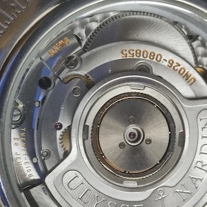 K&ouml;nnte beinhalten: Nahaufnahme eines silbernen Uhrwerks mit komplizierten Zahnr&auml;dern und Komponenten. Die Uhr zeigt den Text "SWISS" und "TWENTY-EIGHT" sowie den Markennamen "ULYSSE NARDIN". Die Modellnummer "UN026-080855" ist ebenfalls sichtbar.