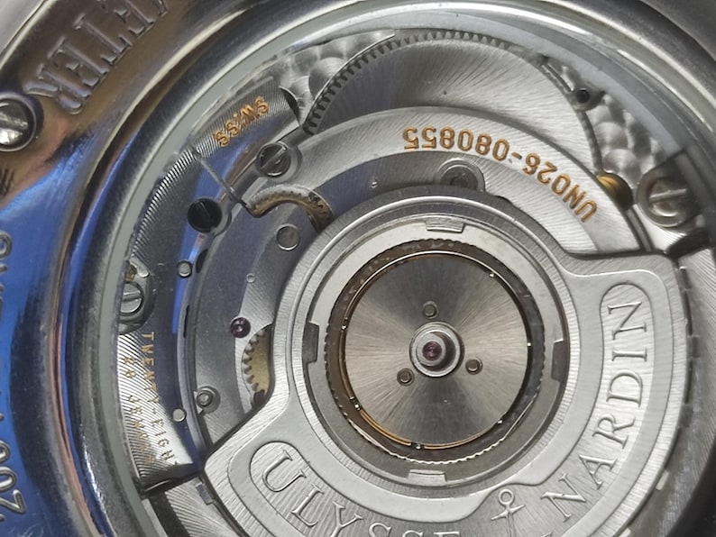 K&ouml;nnte beinhalten: Nahaufnahme eines silbernen Uhrwerks mit komplizierten Zahnr&auml;dern und Komponenten. Die Uhr zeigt den Text "SWISS" und "TWENTY-EIGHT" sowie den Markennamen "ULYSSE NARDIN". Die Modellnummer "UN026-080855" ist ebenfalls sichtbar.