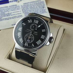 K&ouml;nnte beinhalten: Eine Luxus-Armbanduhr mit schwarzem Zifferblatt, silberner L&uuml;nette und schwarzem Kautschukarmband. Die Uhr hat r&ouml;mische Ziffern und die Marke "ULYSSE NARDIN" ist sichtbar. Die Uhr wird in einer cremefarbenen Box pr&auml;sentiert.