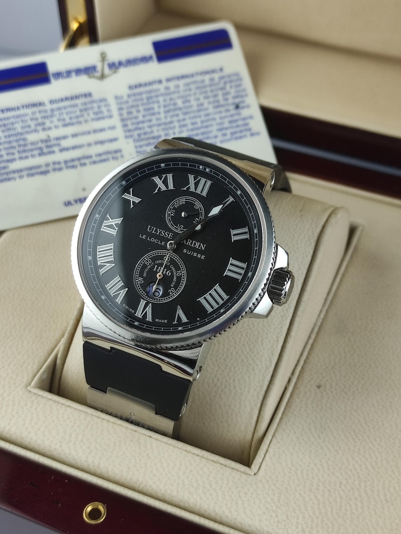 K&ouml;nnte beinhalten: Eine Luxus-Armbanduhr mit schwarzem Zifferblatt, silberner L&uuml;nette und schwarzem Kautschukarmband. Die Uhr hat r&ouml;mische Ziffern und die Marke "ULYSSE NARDIN" ist sichtbar. Die Uhr wird in einer cremefarbenen Box pr&auml;sentiert.
