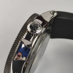 Puede incluir: Primer plano de un reloj de pulsera plateado con una correa negra. El reloj presenta un bisel texturizado, una corona con un logotipo y el n&uacute;mero "263.67" grabado en el lateral. El reloj tiene un dise&ntilde;o elegante y moderno.