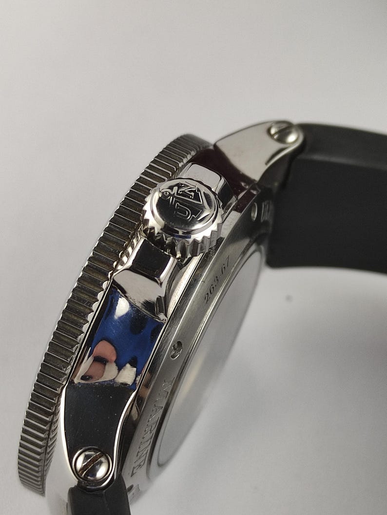 Puede incluir: Primer plano de un reloj de pulsera plateado con una correa negra. El reloj presenta un bisel texturizado, una corona con un logotipo y el n&uacute;mero "263.67" grabado en el lateral. El reloj tiene un dise&ntilde;o elegante y moderno.