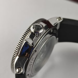 K&ouml;nnte beinhalten: Nahaufnahme einer silberfarbenen Uhr mit schwarzem Armband. Die Uhr hat eine strukturierte L&uuml;nette und eine Krone mit Logo. Das Wort "MARINE" ist auf dem Zifferblatt zu sehen. Es handelt sich um einen Luxusartikel.