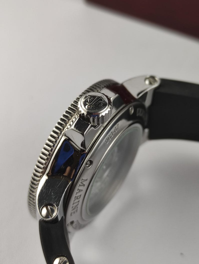 K&ouml;nnte beinhalten: Nahaufnahme einer silberfarbenen Uhr mit schwarzem Armband. Die Uhr hat eine strukturierte L&uuml;nette und eine Krone mit Logo. Das Wort "MARINE" ist auf dem Zifferblatt zu sehen. Es handelt sich um einen Luxusartikel.