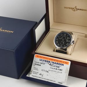 Puede incluir: Un reloj Ulysse Nardin con esfera negra y caja plateada, exhibido en una caja de madera. El reloj tiene n&uacute;meros romanos y una correa de cuero negro. Tambi&eacute;n se ven una caja azul y una tarjeta de garant&iacute;a.