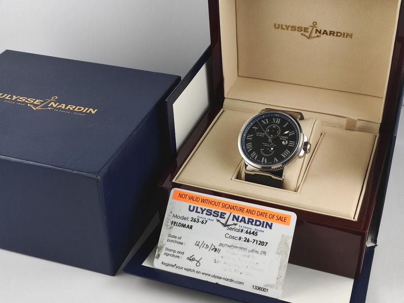 Puede incluir: Un reloj Ulysse Nardin con esfera negra y caja plateada, exhibido en una caja de madera. El reloj tiene n&uacute;meros romanos y una correa de cuero negro. Tambi&eacute;n se ven una caja azul y una tarjeta de garant&iacute;a.