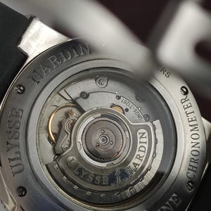 Puede incluir: Primer plano de la parte trasera de un reloj Ulysse Nardin Marine Chronometer. El mecanismo del reloj es visible a trav&eacute;s de una parte trasera transparente. El reloj tiene una correa negra y una caja plateada con el nombre de la marca grabado.