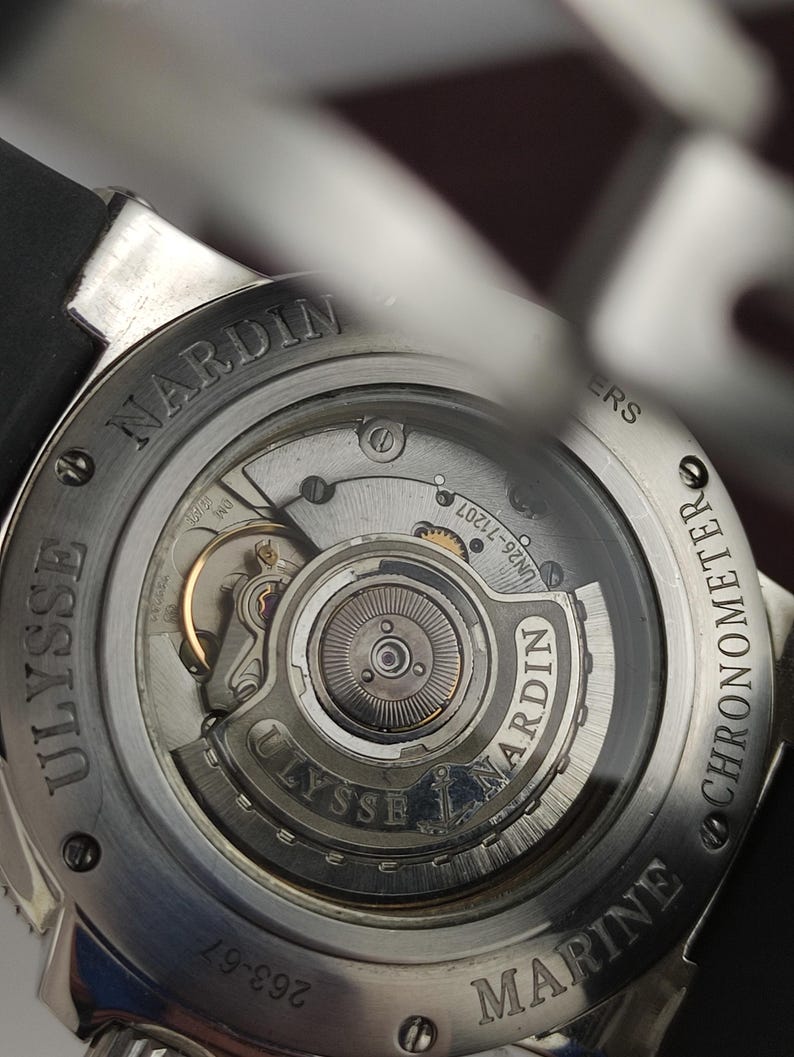 Puede incluir: Primer plano de la parte trasera de un reloj Ulysse Nardin Marine Chronometer. El mecanismo del reloj es visible a trav&eacute;s de una parte trasera transparente. El reloj tiene una correa negra y una caja plateada con el nombre de la marca grabado.