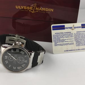 K&ouml;nnte beinhalten: Eine Ulysse Nardin Uhr mit schwarzem Zifferblatt, silberfarbenem Geh&auml;use und schwarzem Kautschukarmband. Die Uhr verf&uuml;gt &uuml;ber r&ouml;mische Ziffern und Hilfszifferbl&auml;tter. Eine Garantiekarte und eine Holzbox mit dem Markenlogo sind ebenfalls vorhanden.