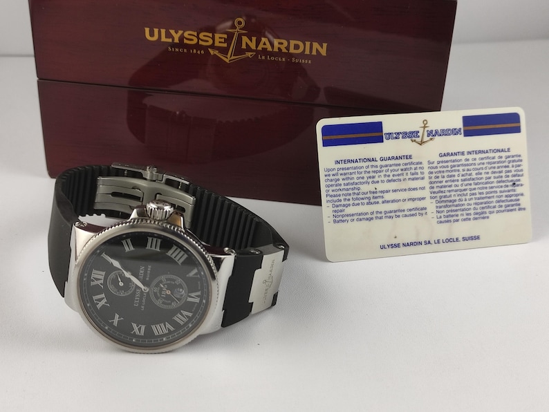 K&ouml;nnte beinhalten: Eine Ulysse Nardin Uhr mit schwarzem Zifferblatt, silberfarbenem Geh&auml;use und schwarzem Kautschukarmband. Die Uhr verf&uuml;gt &uuml;ber r&ouml;mische Ziffern und Hilfszifferbl&auml;tter. Eine Garantiekarte und eine Holzbox mit dem Markenlogo sind ebenfalls vorhanden.