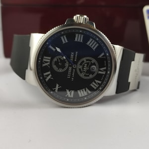 K&ouml;nnte beinhalten: Eine silberfarbene Armbanduhr mit schwarzem Zifferblatt und schwarzem Kautschukarmband. Das Zifferblatt zeigt r&ouml;mische Ziffern und zwei kleinere Zifferbl&auml;tter. Der Markenname "Ulysse Nardin" ist sichtbar.