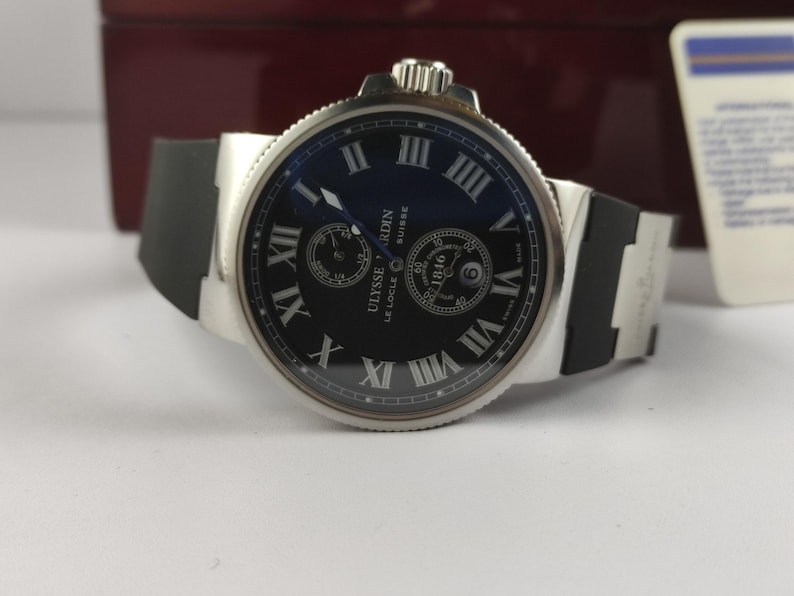 K&ouml;nnte beinhalten: Eine silberfarbene Armbanduhr mit schwarzem Zifferblatt und schwarzem Kautschukarmband. Das Zifferblatt zeigt r&ouml;mische Ziffern und zwei kleinere Zifferbl&auml;tter. Der Markenname "Ulysse Nardin" ist sichtbar.