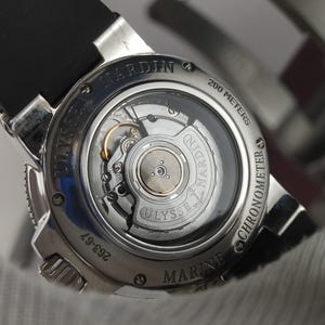 K&ouml;nnte beinhalten: Nahaufnahme einer silbernen Ulysse Nardin Marine Chronometer Uhr. Die Uhr hat ein schwarzes Armband und einen transparenten R&uuml;cken, der die inneren Mechanismen zeigt. Wasserdicht bis 200 Meter.