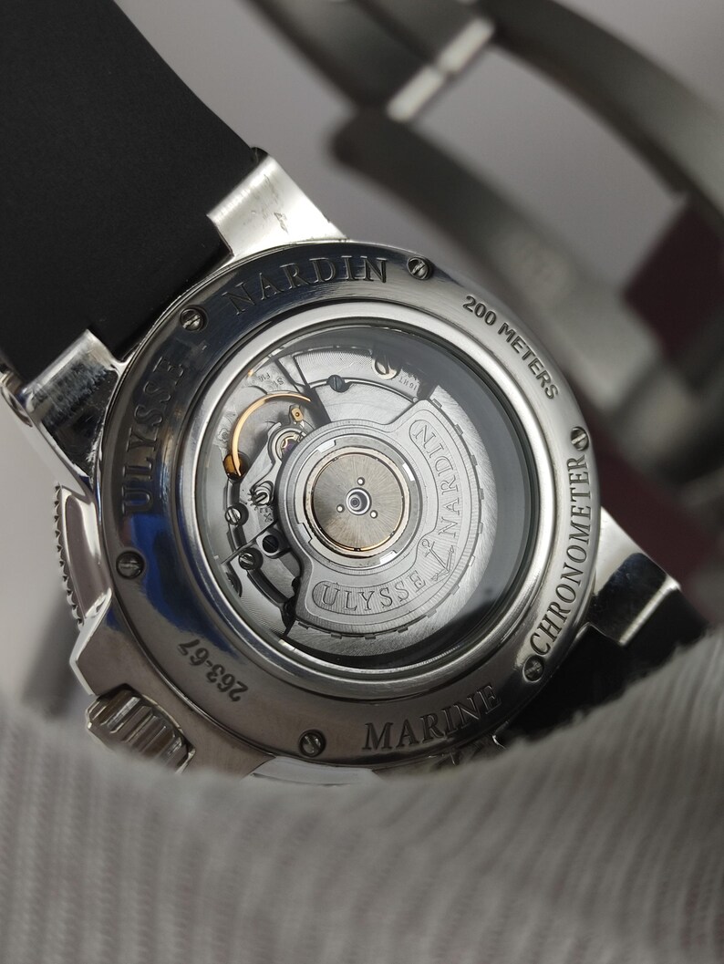 K&ouml;nnte beinhalten: Nahaufnahme einer silbernen Ulysse Nardin Marine Chronometer Uhr. Die Uhr hat ein schwarzes Armband und einen transparenten R&uuml;cken, der die inneren Mechanismen zeigt. Wasserdicht bis 200 Meter.