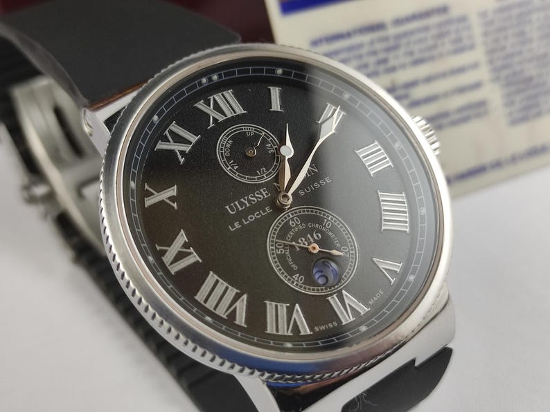 K&ouml;nnte beinhalten: Nahaufnahme einer Luxus-Armbanduhr mit schwarzem Zifferblatt und silbernem Geh&auml;use. Die Uhr hat r&ouml;mische Ziffern und zwei Hilfszifferbl&auml;tter. Sie hat ein schwarzes Lederarmband. Der Markenname "ULYSSE NARDIN" ist sichtbar.