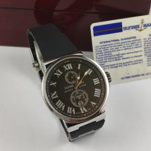 K&ouml;nnte beinhalten: Eine luxuri&ouml;se Ulysse Nardin Uhr mit schwarzem Lederarmband. Das dunkle Zifferblatt zeigt r&ouml;mische Ziffern und ein kleines Hilfszifferblatt. Die Uhr wird mit ihrer Box und Garantiekarte pr&auml;sentiert.