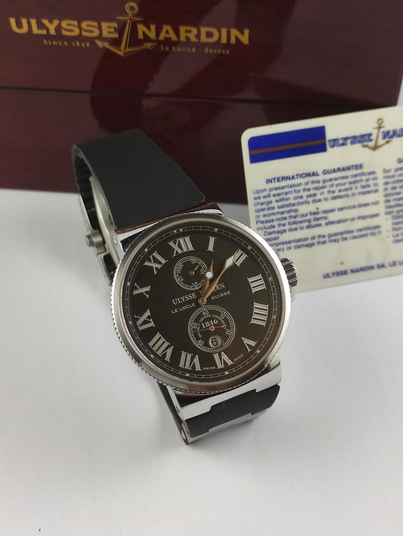 K&ouml;nnte beinhalten: Eine luxuri&ouml;se Ulysse Nardin Uhr mit schwarzem Lederarmband. Das dunkle Zifferblatt zeigt r&ouml;mische Ziffern und ein kleines Hilfszifferblatt. Die Uhr wird mit ihrer Box und Garantiekarte pr&auml;sentiert.