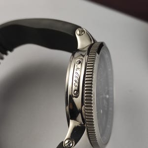 Puede incluir: Primer plano de un reloj de pulsera plateado con esfera negra y correa de caucho negra. El reloj tiene un bisel texturizado y el texto "NCC43" es visible en el lateral.