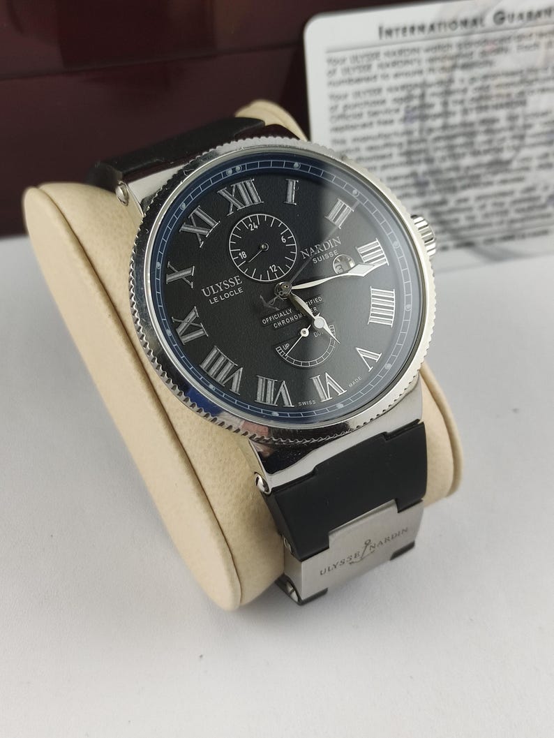Puede incluir: Un reloj de lujo con esfera negra, bisel plateado y correa de caucho negra. El reloj presenta n&uacute;meros romanos y subesferas. La marca "Ulysse Nardin" es visible en la esfera y la correa. El reloj se exhibe sobre un coj&iacute;n color crema.