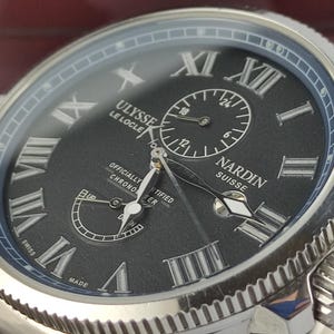 Puede incluir: Primer plano de un reloj de lujo con esfera negra y caja plateada. El reloj presenta n&uacute;meros romanos, subesferas y el texto "ULYSSE LE LOCLE NARDIN SUISSE". Es un cron&oacute;metro certificado.
