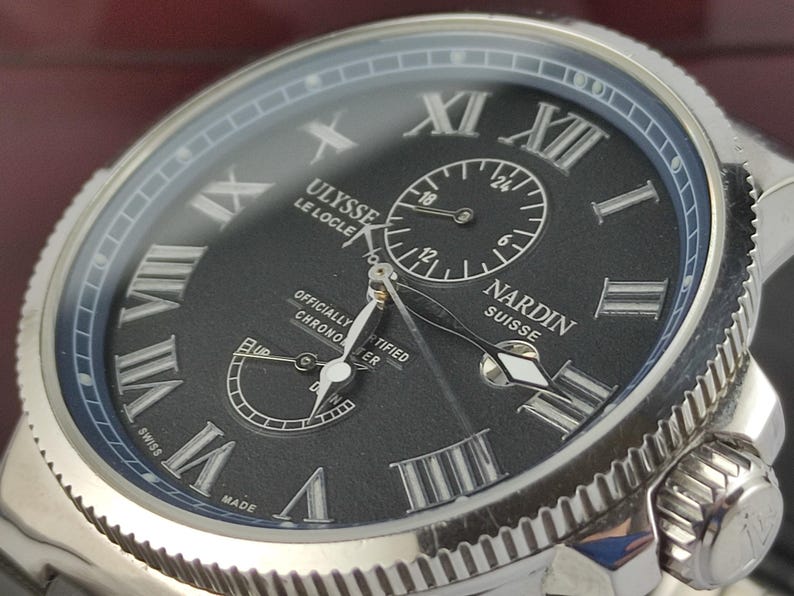 Puede incluir: Primer plano de un reloj de lujo con esfera negra y caja plateada. El reloj presenta n&uacute;meros romanos, subesferas y el texto "ULYSSE LE LOCLE NARDIN SUISSE". Es un cron&oacute;metro certificado.