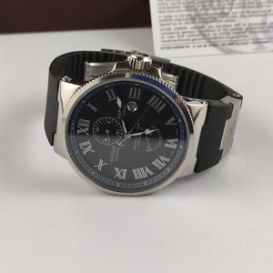 Puede incluir: Un reloj de pulsera plateado con esfera negra y correa de caucho negra. La esfera del reloj presenta n&uacute;meros romanos y dos esferas m&aacute;s peque&ntilde;as. El reloj est&aacute; sobre una superficie blanca.