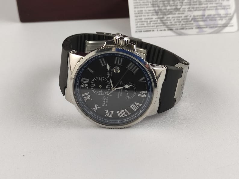Puede incluir: Un reloj de pulsera plateado con esfera negra y correa de caucho negra. La esfera del reloj presenta n&uacute;meros romanos y dos esferas m&aacute;s peque&ntilde;as. El reloj est&aacute; sobre una superficie blanca.