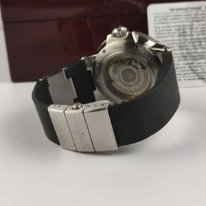 Puede incluir: Un reloj de lujo con una correa de caucho negro y un cierre plateado. La esfera del reloj es visible, mostrando intrincados mecanismos internos. El reloj est&aacute; sobre una superficie blanca, con una caja roja oscura en el fondo.