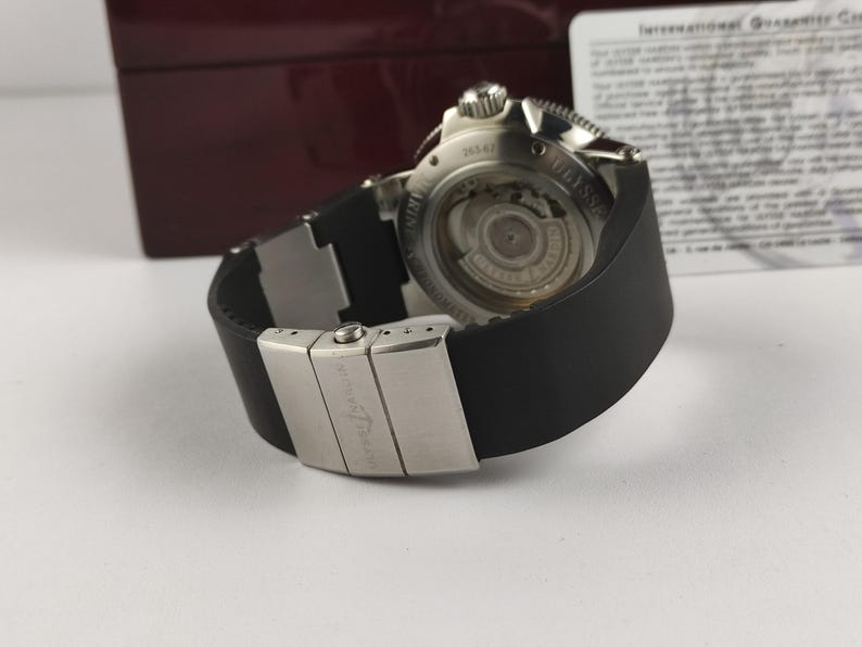 Puede incluir: Un reloj de lujo con una correa de caucho negro y un cierre plateado. La esfera del reloj es visible, mostrando intrincados mecanismos internos. El reloj est&aacute; sobre una superficie blanca, con una caja roja oscura en el fondo.