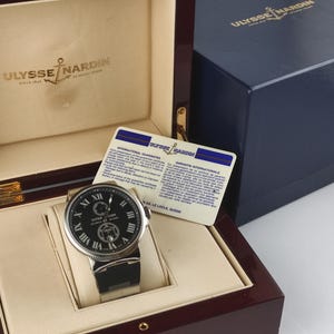 K&ouml;nnte beinhalten: Eine luxuri&ouml;se Ulysse Nardin Uhr mit schwarzem Zifferblatt und silberfarbenem Geh&auml;use, pr&auml;sentiert in einer Holzbox. Die Uhr hat r&ouml;mische Ziffern und ein schwarzes Armband. Eine blaue Box und eine Garantiekarte sind ebenfalls vorhanden.