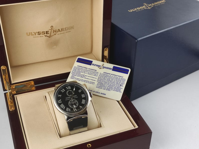 K&ouml;nnte beinhalten: Eine luxuri&ouml;se Ulysse Nardin Uhr mit schwarzem Zifferblatt und silberfarbenem Geh&auml;use, pr&auml;sentiert in einer Holzbox. Die Uhr hat r&ouml;mische Ziffern und ein schwarzes Armband. Eine blaue Box und eine Garantiekarte sind ebenfalls vorhanden.