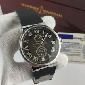 K&ouml;nnte beinhalten: Eine luxuri&ouml;se Armbanduhr mit schwarzem Zifferblatt, silberner L&uuml;nette und schwarzem Kautschukarmband. Die Uhr verf&uuml;gt &uuml;ber r&ouml;mische Ziffern und Hilfszifferbl&auml;tter. Der Markenname "Ulysse Nardin" ist auf dem Zifferblatt und der Box zu sehen.